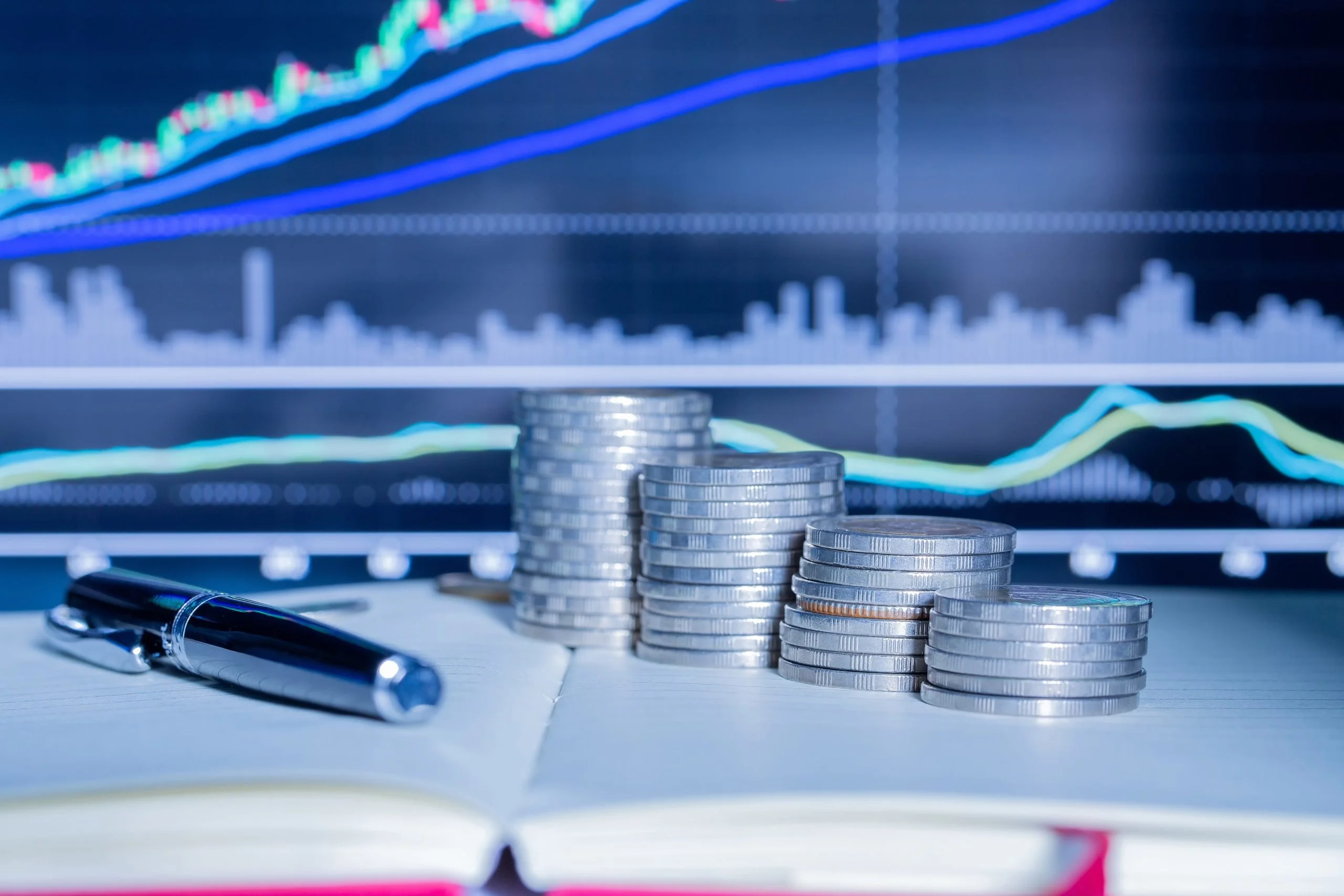 Brokers trading: monedas y un boligrafo sobre un libro y con gráficas de fondo.